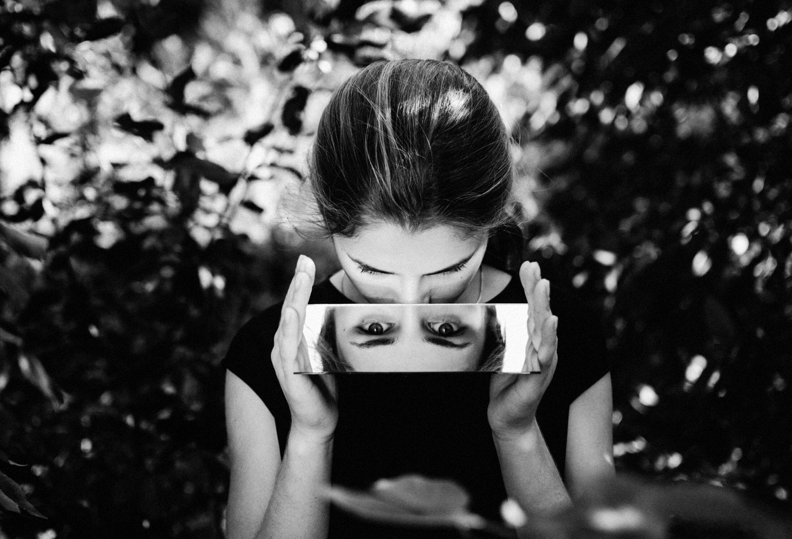 Black and white portrait of a woman gazing into a mirror outdoors, capturing reflection.